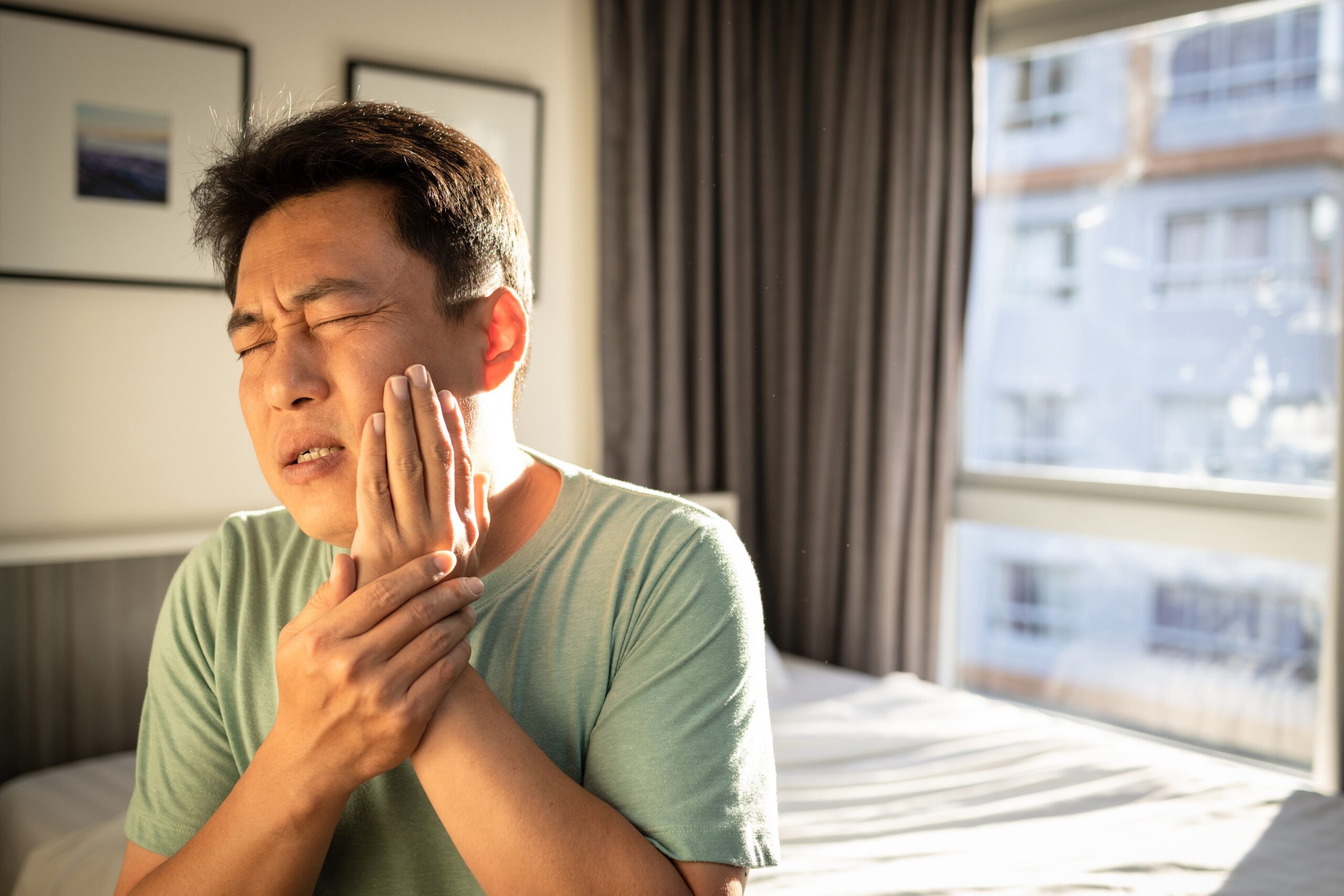 man sitting up in bed with his hand over his jaw, extreme pain and possible TMJ